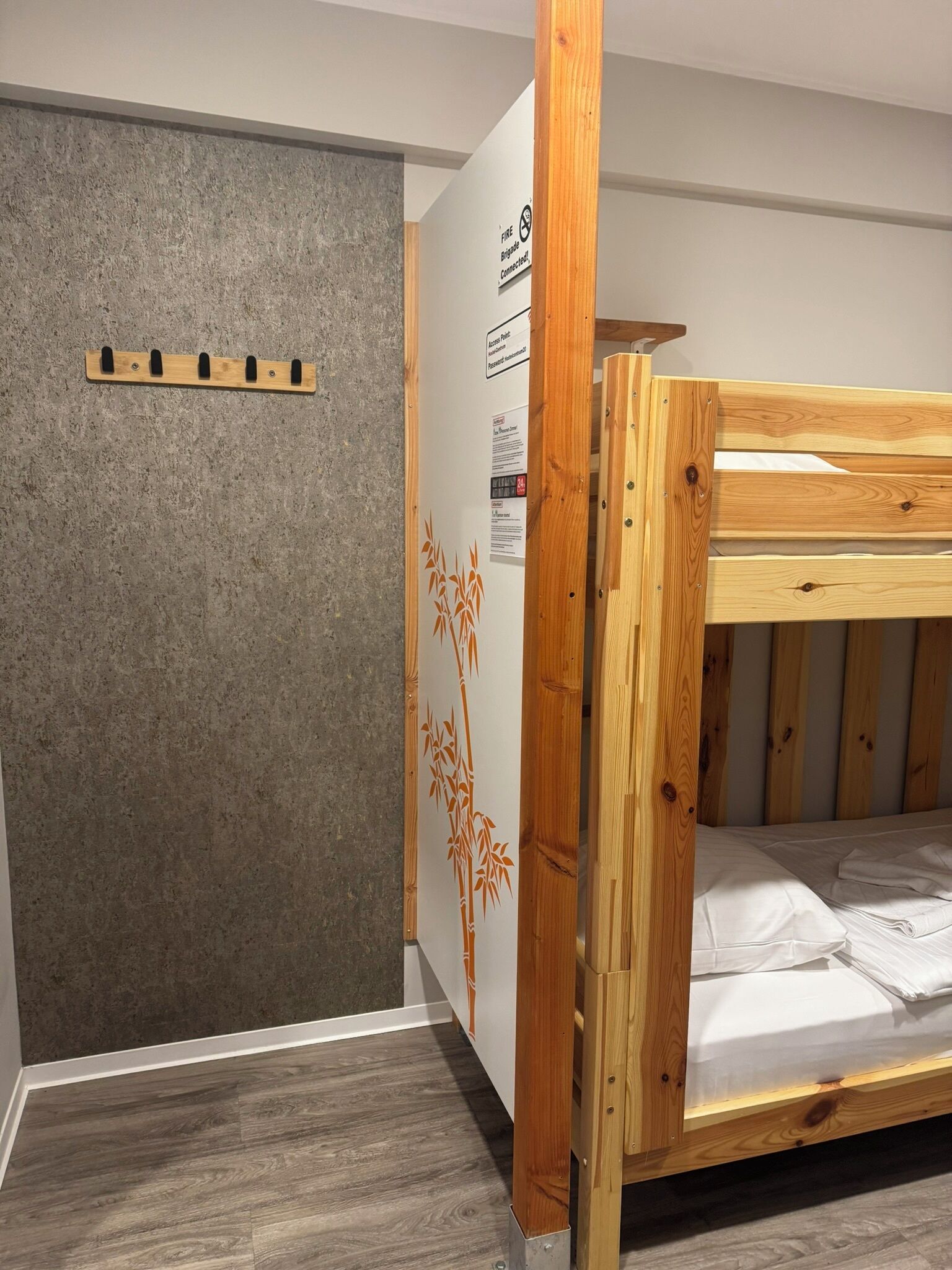 Una cama litera con estructura de madera y ropa de cama blanca, una pared gris con textura y una puerta decorada con motivos de bambú.
