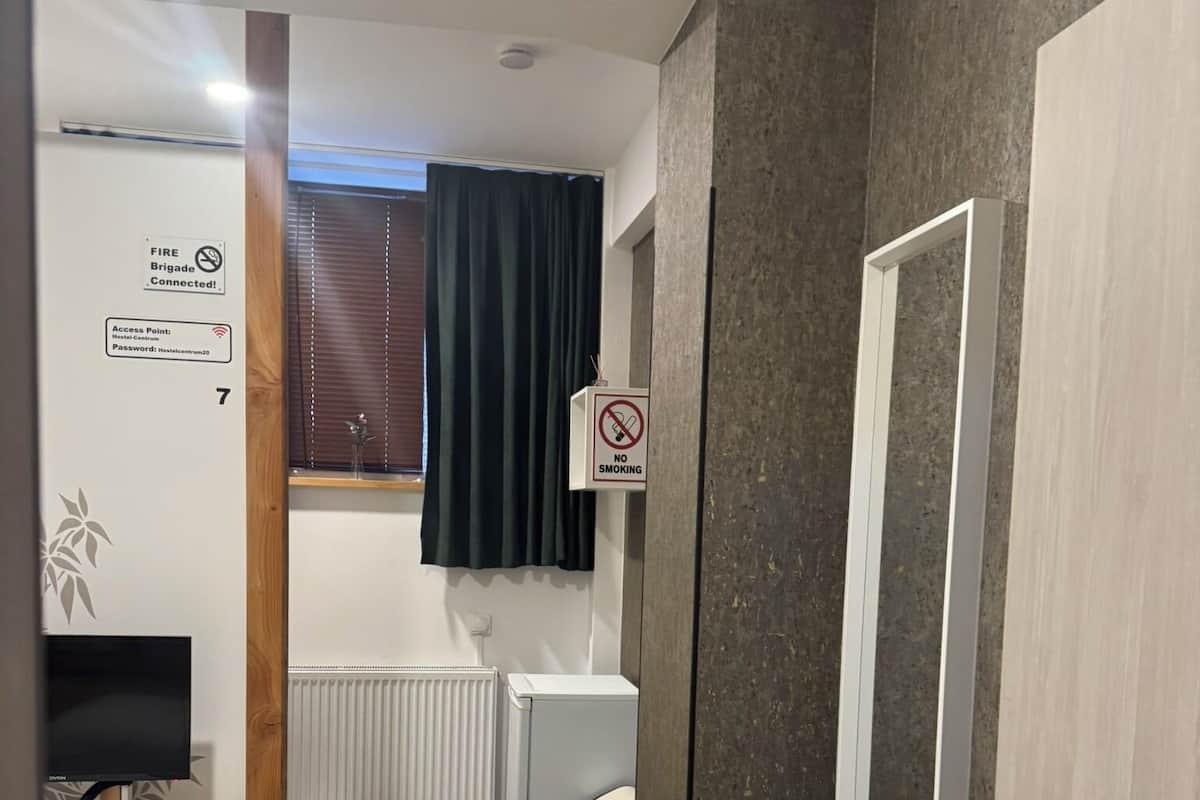 A hotel room with a bed, a desk with a computer, a radiator, a trash bin, and a door with a security lock.