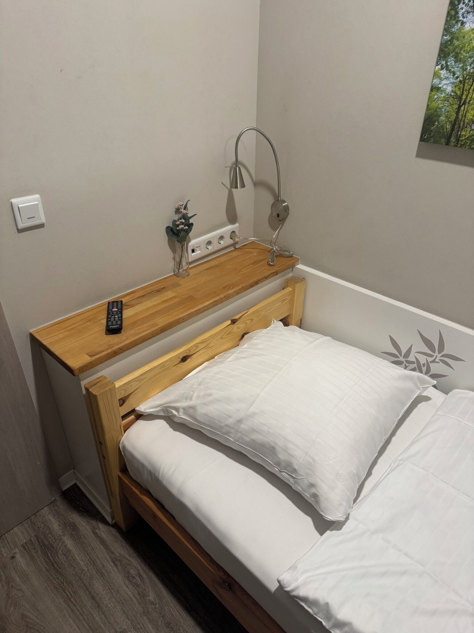 A small, single-bed room with a wooden headboard, a bedside table with a lamp, a power outlet, and a remote control on the table.