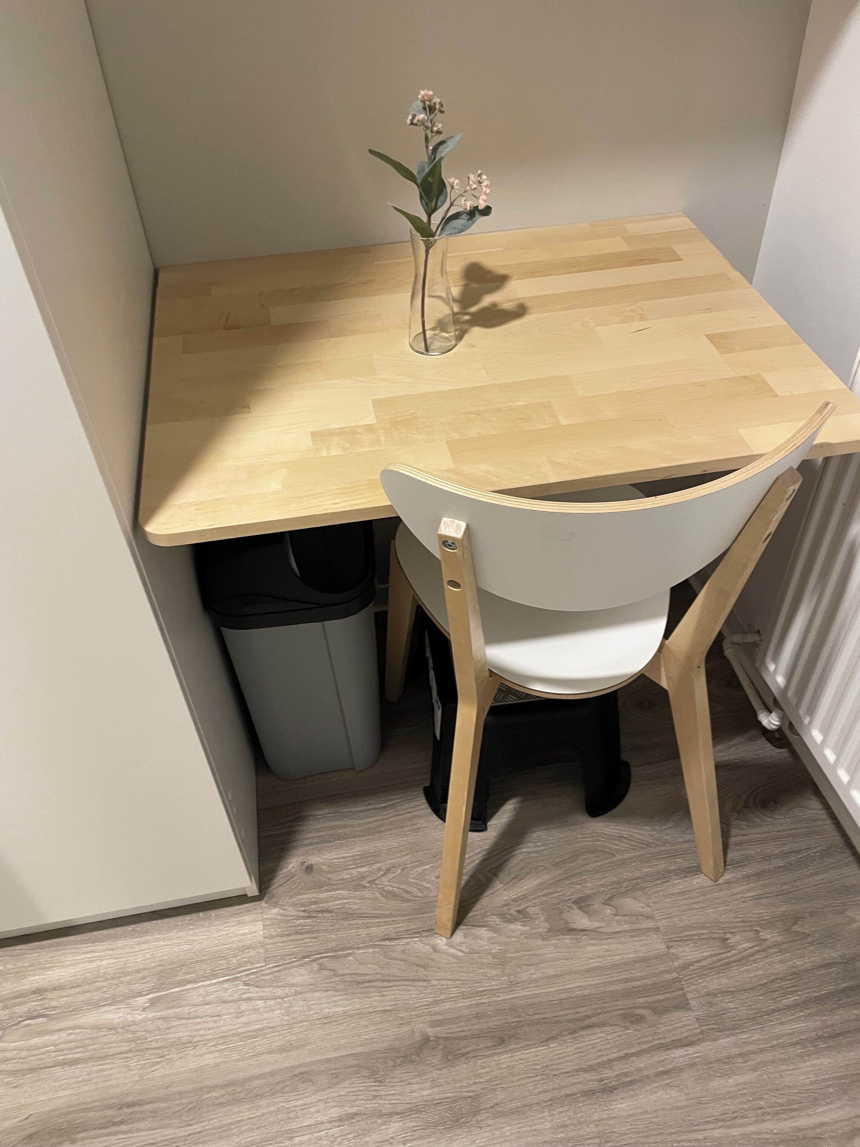 Ein Eckzimmer mit Holztisch, einem weißen Stuhl, einem Mülleimer und einer Heizung.