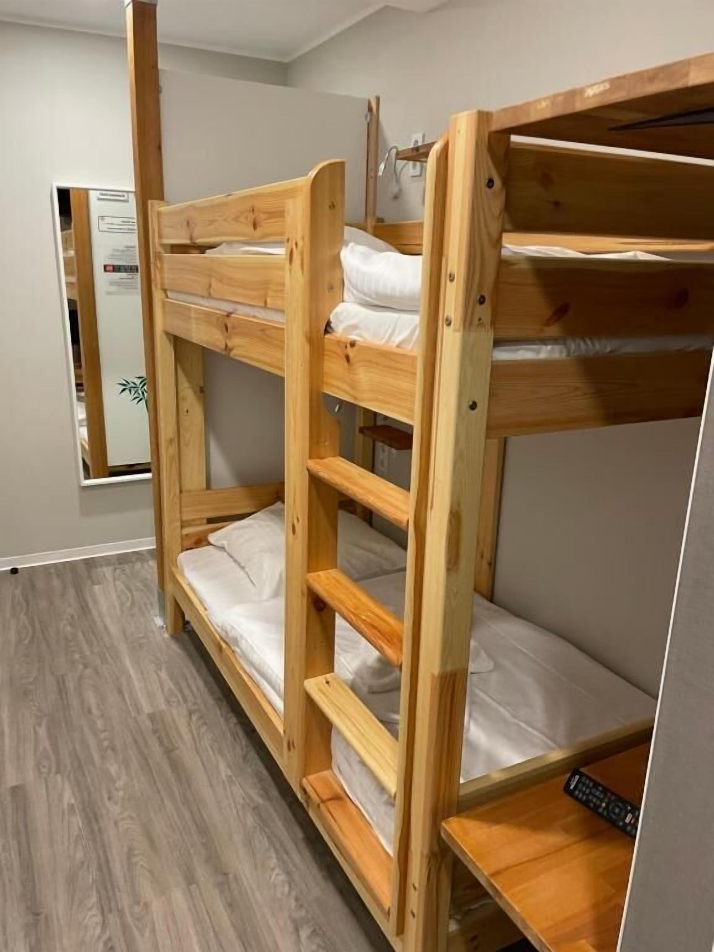 Una cama litera con estructura de madera y ropa de cama blanca, una mesita de noche de madera y un espejo en la pared.