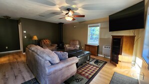 Un salon comprenant un canapé, un fauteuil, un pouf, un ventilateur de plafond, un téléviseur et une fenêtre donnant sur des arbres.