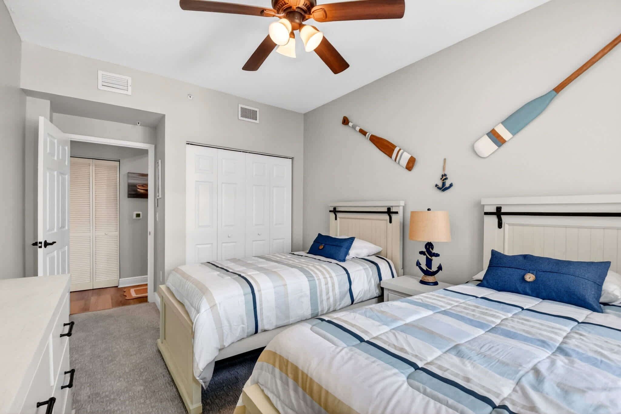 A bedroom with two beds, a ceiling fan, a nightstand, a lamp, and nautical-themed wall decor.