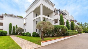 Een modern gebouw met een witte gevel, meerdere balkons en een verzorgde tuin met verschillende struiken en bomen.