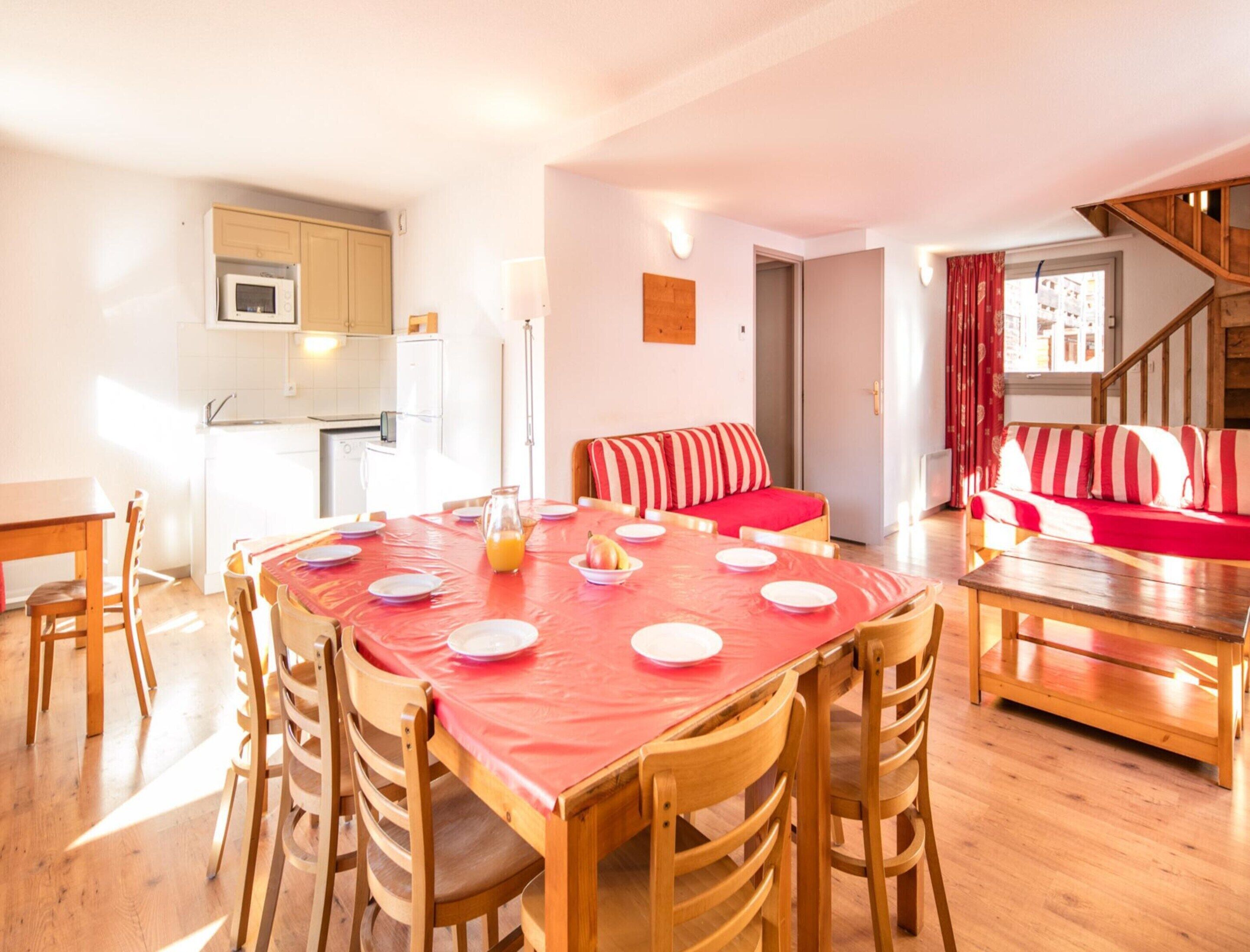 A dining area with a table set for four, a kitchen in the background, and a staircase leading to another level.