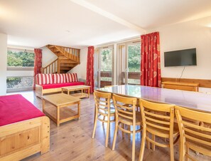 A room with a wooden staircase, a sofa with red cushions, a dining table with chairs, and a TV mounted on the wall. - Duplex for 12 Near Galibier Pass (Valloire)