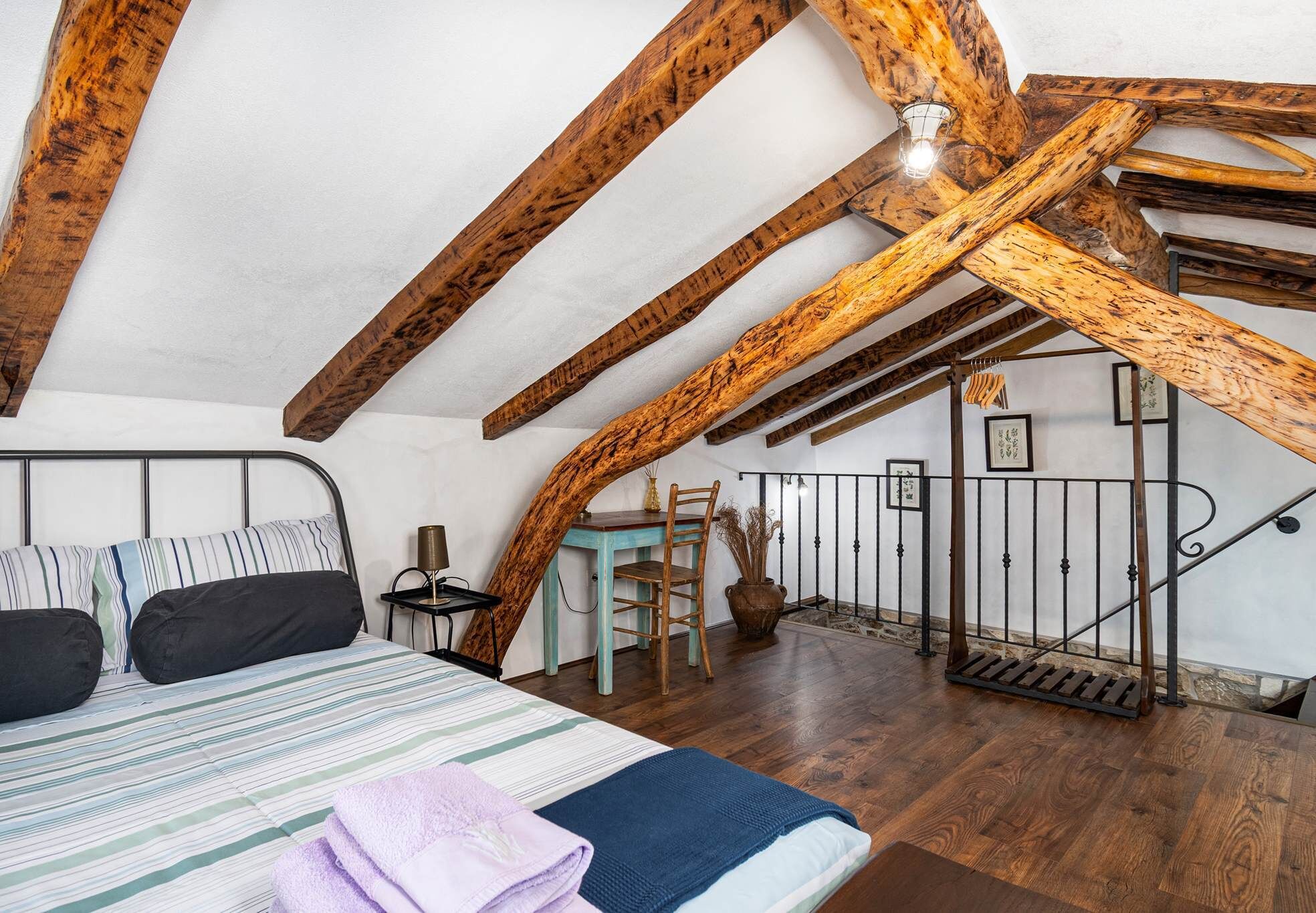 A bedroom with a wooden ceiling, a bed with striped bedding, a chair, a small table, and a staircase.