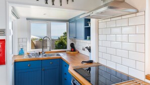 A modern kitchen with blue cabinets, a stainless steel sink, and a view of the sea. - Red Cedars (Bembridge)