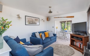 A living room with a blue sofa, a television, a bookshelf, and a dining area with white chairs.