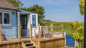 A small, light blue cottage with a wooden deck, a covered outdoor seating area, and a view of a grassy field. - Red Cedars (Bembridge)