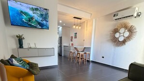 A modern living room with a flat-screen TV, a sofa, a dining area with stools, and a kitchen in the background.