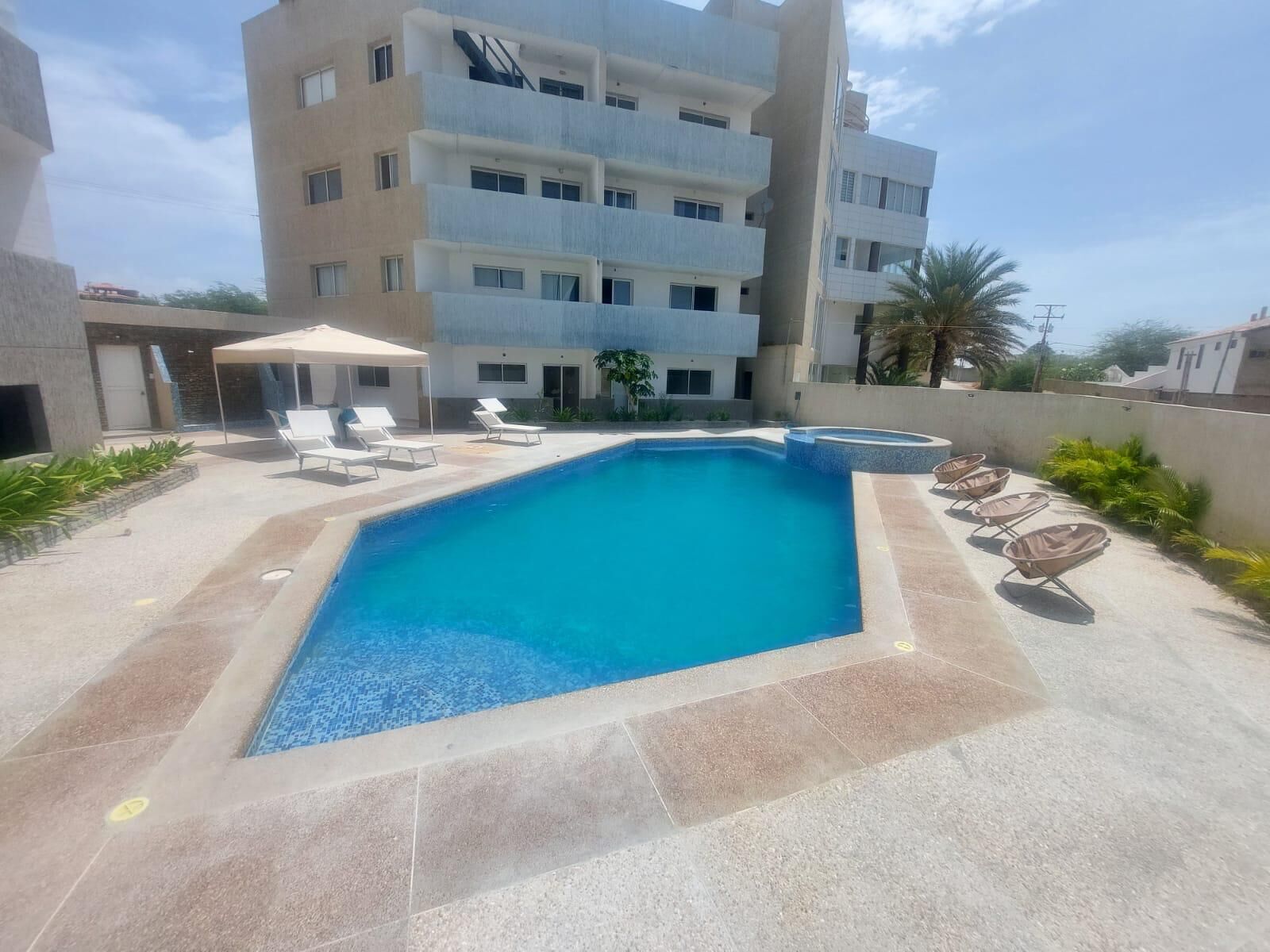 A swimming pool with lounge chairs, a covered seating area, and a multi-story building in the background.