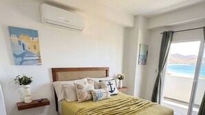 A hotel room with a bed, a nightstand, a wall-mounted air conditioner, a painting, and a view of the beach through a window.