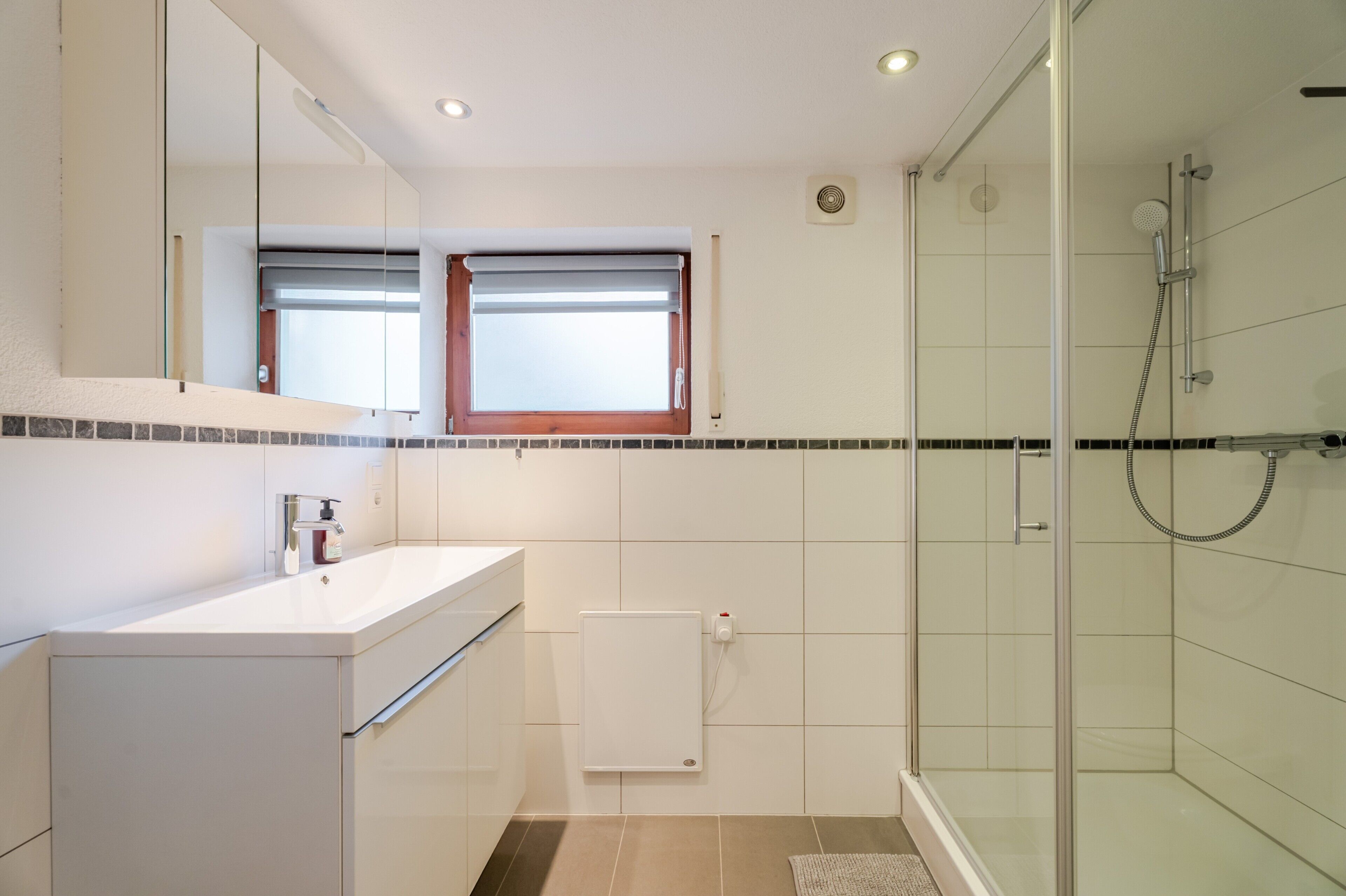 Ein modernes Badezimmer mit einer Duschkabine aus Glas, einem weißen Waschbecken mit Seifenspender und einem Spiegel darüber.