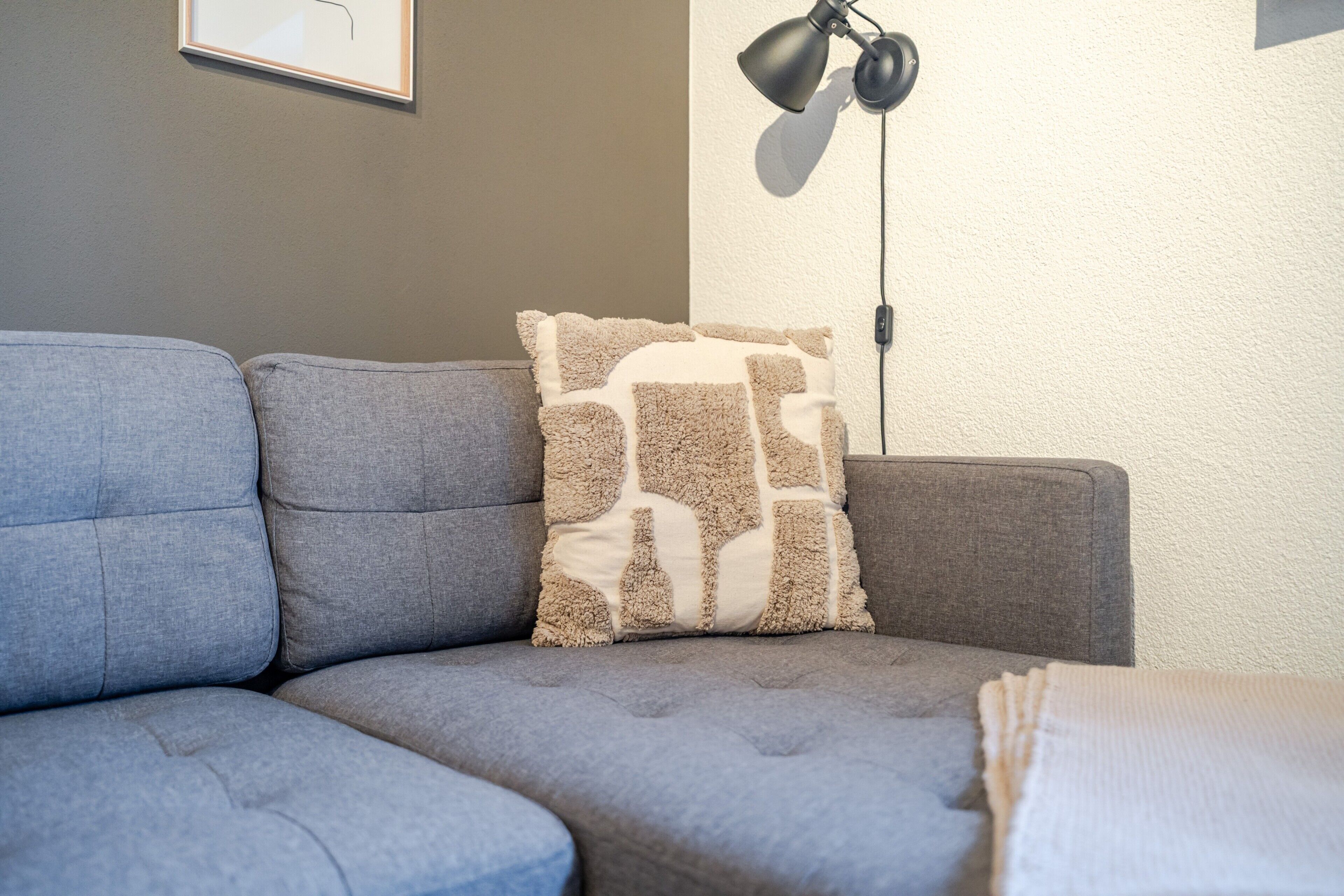 Un coin de pièce avec un canapé gris, un coussin décoratif à motifs et un lampadaire noir.