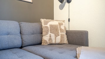 Ein Eckbereich eines Zimmers mit einem grauen Sofa, einem gemusterten Zierkissen und einer schwarzen Stehlampe.