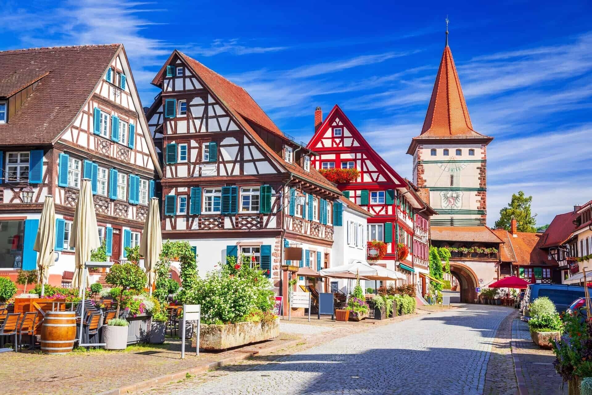 A cobblestone street lined with traditional half-timbered buildings, a prominent clock tower, and outdoor seating areas with plants.