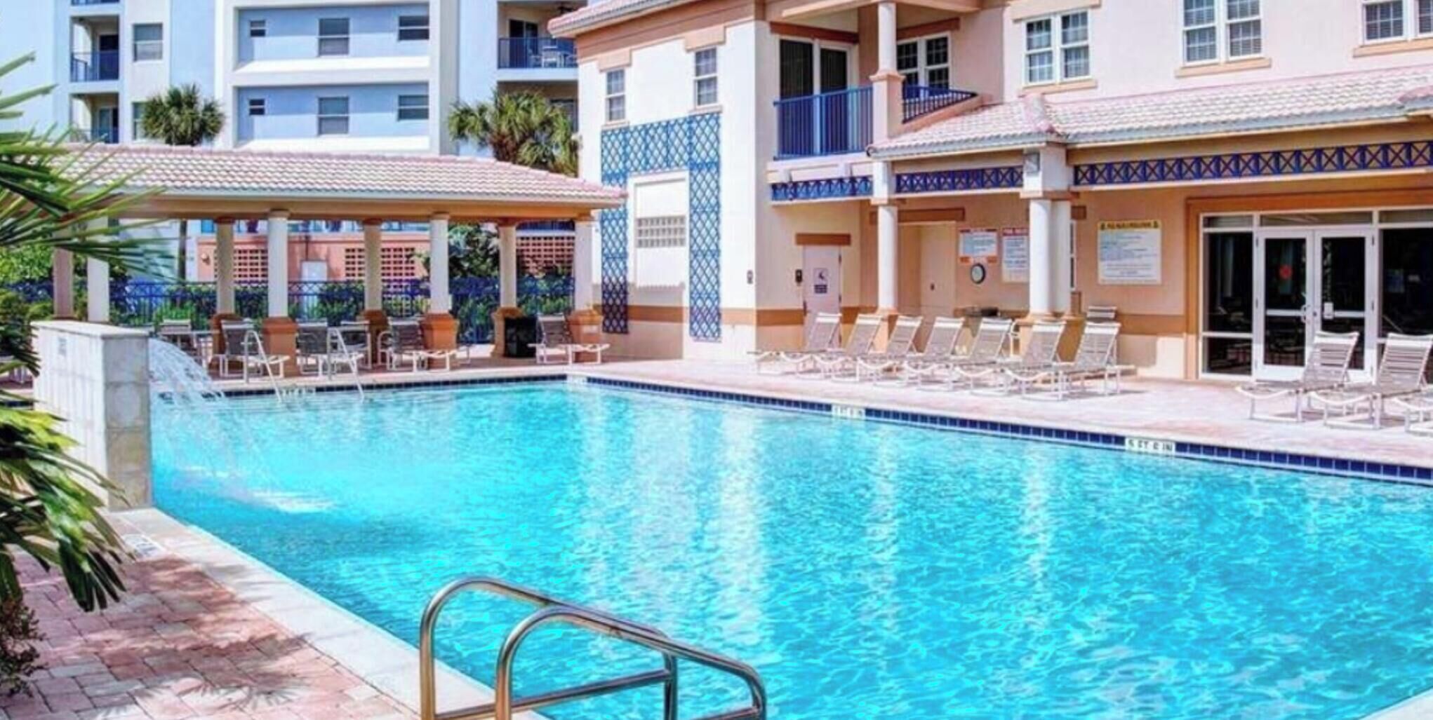 A swimming pool with lounge chairs and a covered seating area, surrounded by multi-story buildings with balconies.