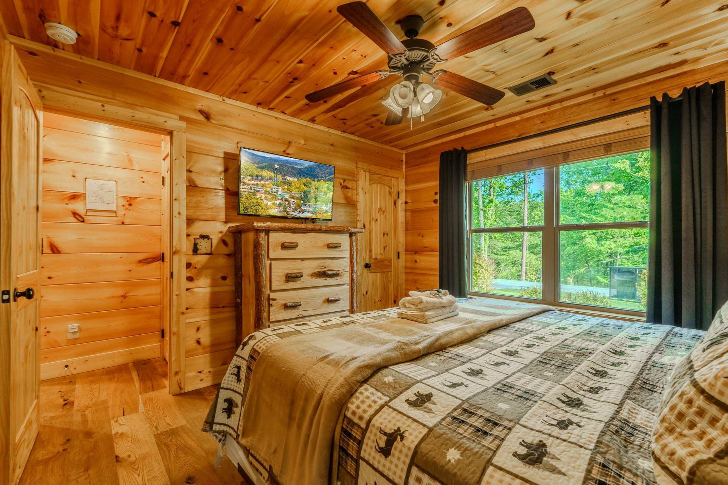 A wooden cabin bedroom with a bed, a ceiling fan, a television, and a window with a view of greenery.