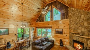 A log cabin interior with a fireplace, dining area, and a large window overlooking greenery. - Gatlinburg-2brs-hot Tub-arcade Games-fireplace (Gatlinburg)