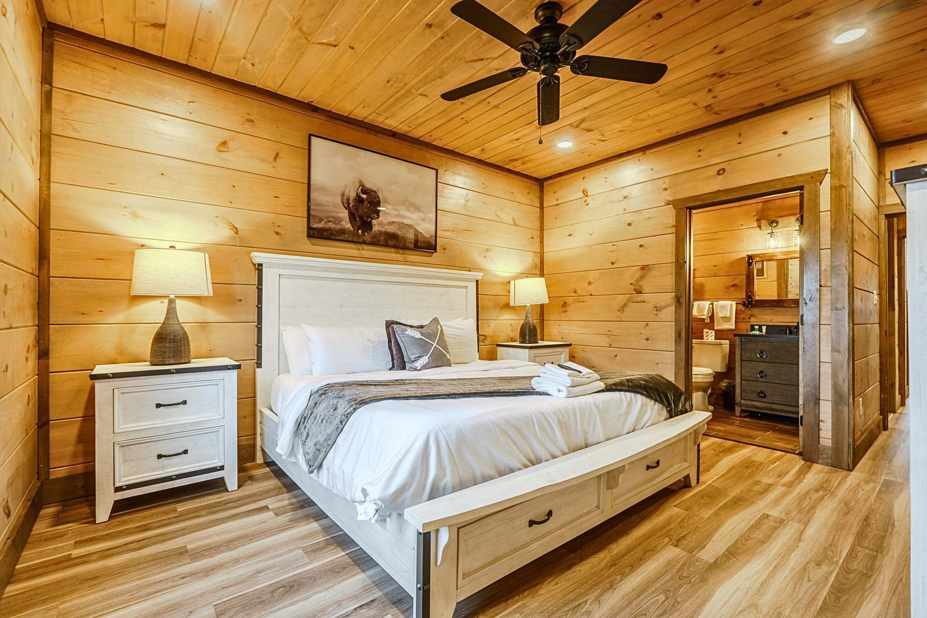 Un dormitorio con paneles de madera, una cama, mesitas de noche, un ventilador de techo y un cuadro.