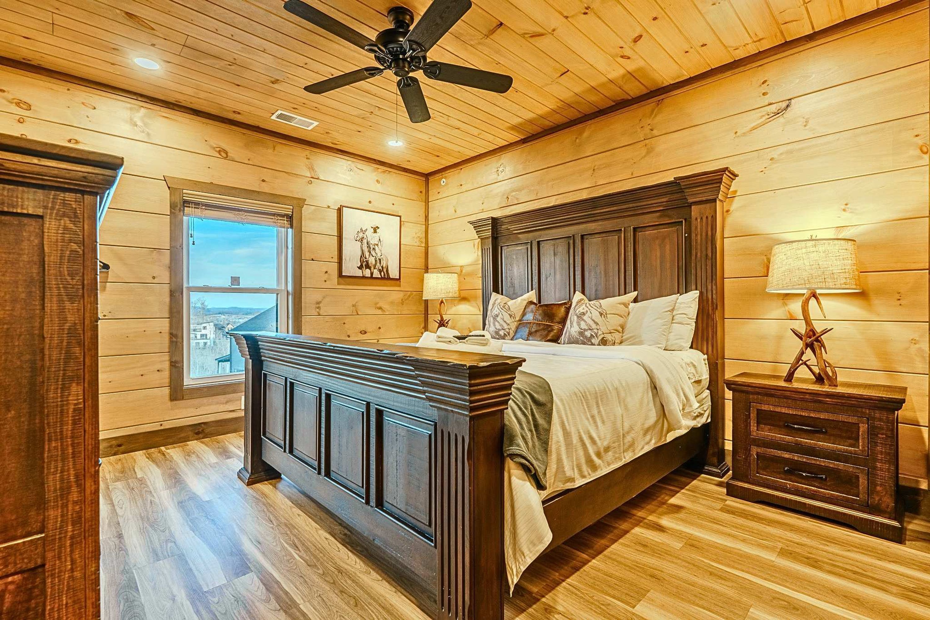 A wooden-paneled bedroom with a large bed, bedside tables, a ceiling fan, and a window with a view.