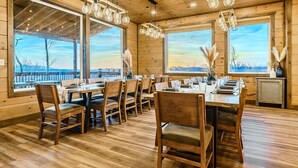 Interior de madera con amplios ventanales, una zona de comedor con mesas y sillas, y lámparas colgantes.