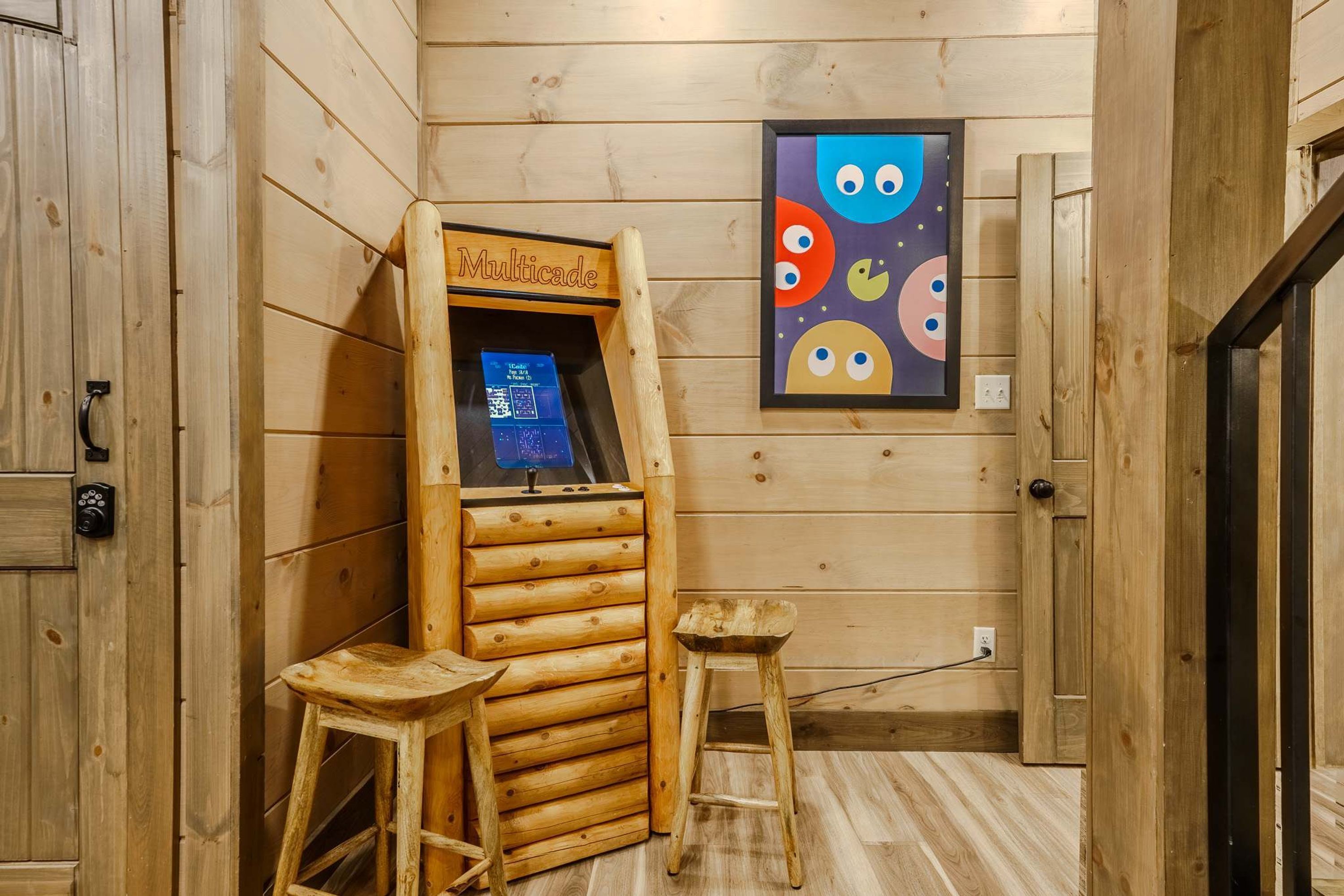 A wooden cabin interior with a Multicade gaming machine, two stools, and a colorful artwork on the wall.