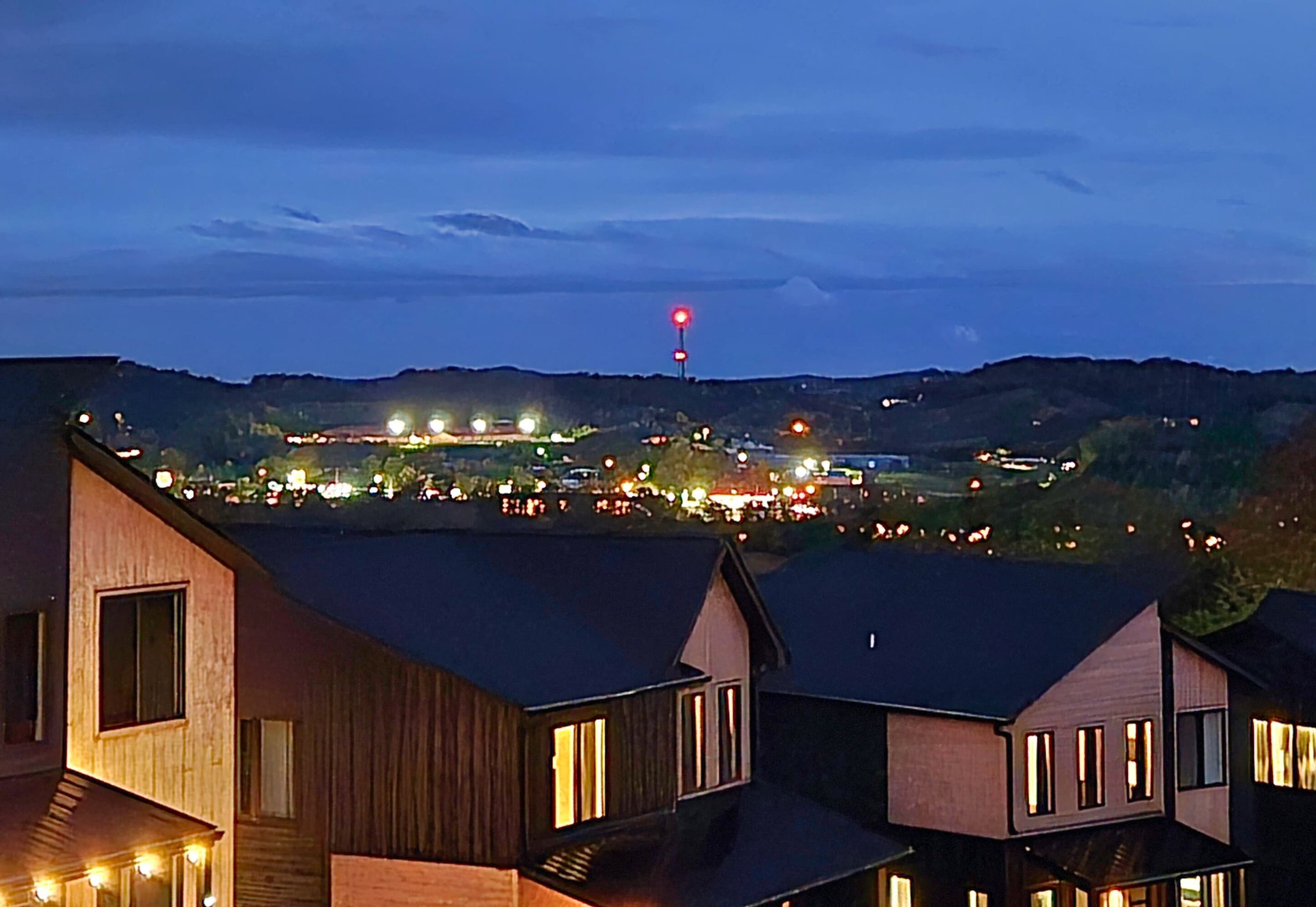 A residential area with modern houses, a prominent red light on a pole, and distant lights indicating a populated area.