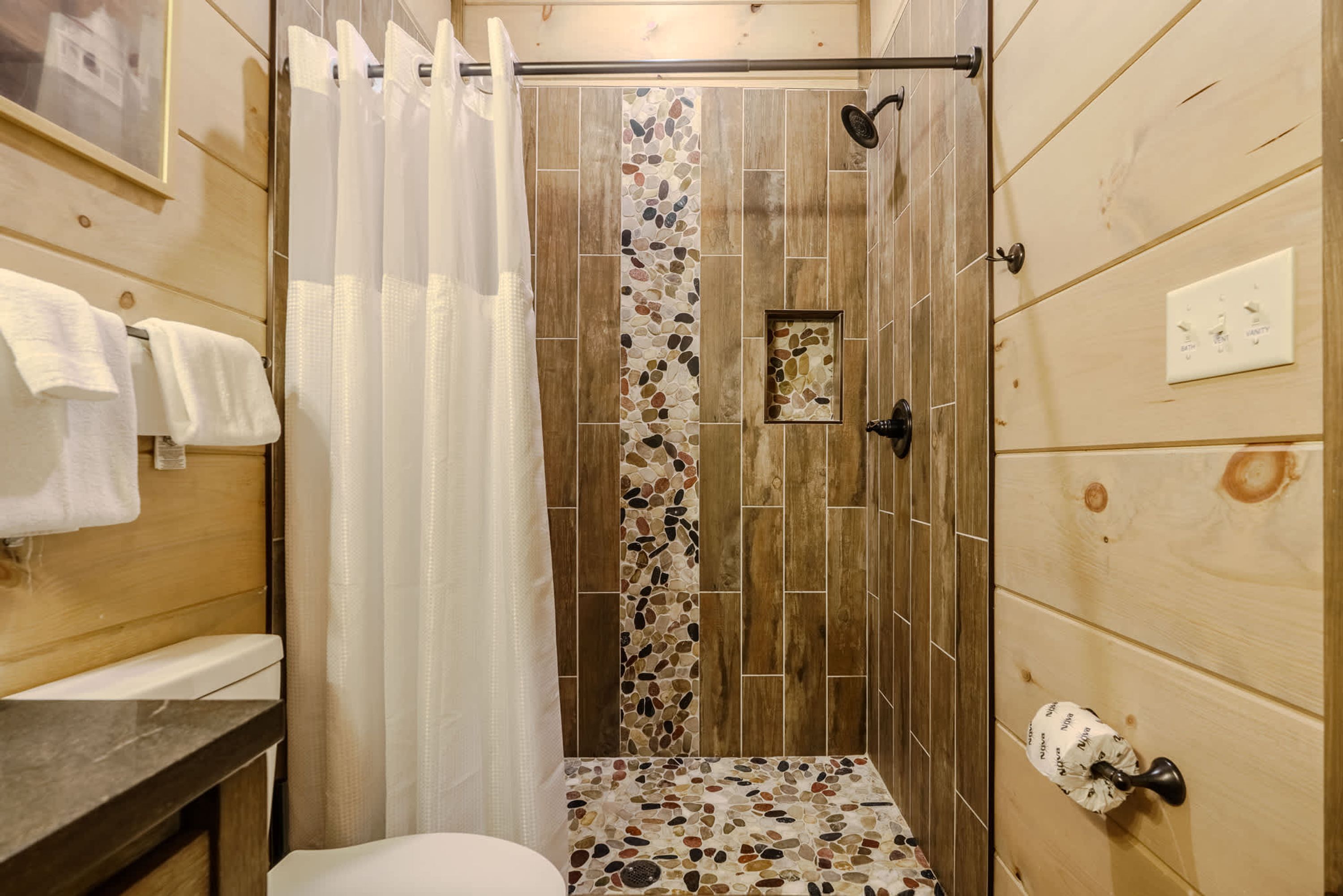 Ducha con pared de madera y suelo de piedras, inodoro blanco y perchero con toallas.