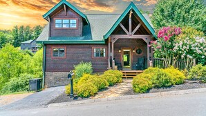 A wooden cabin with a green roof, a porch, and a garden with pink flowers. - NEW Lux 4 BR Pigeon Forge Dollywood Hot Tub (Pigeon Forge)