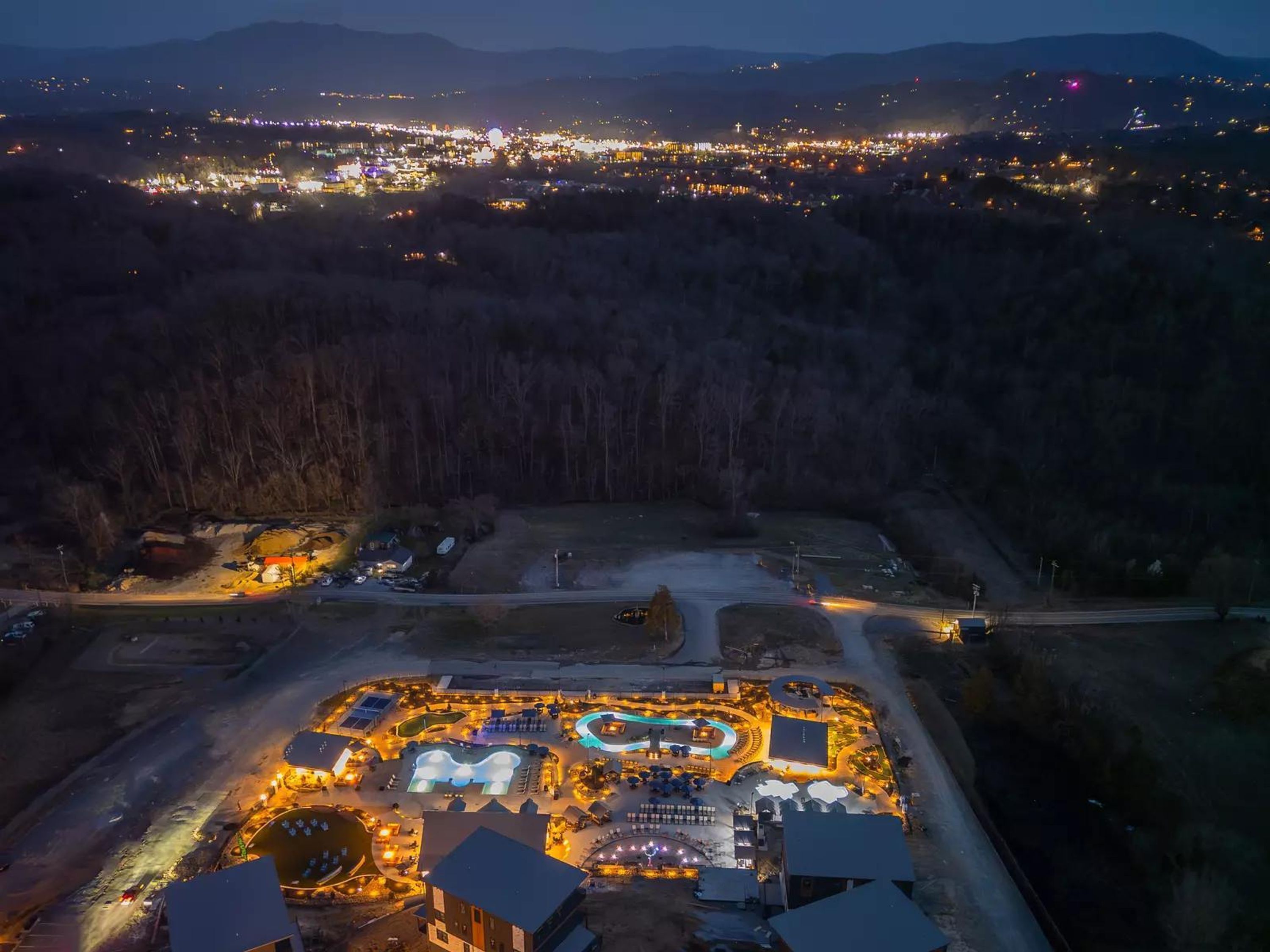 An aerial view of a resort with a central pool area, multiple buildings, and a surrounding forested area.