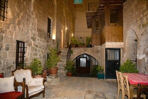 A stone courtyard with a wooden bench, a table with a red tablecloth, and potted plants. - NAHIL MİSAFİREVİ MARDİN (ARTUKLU)