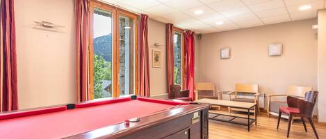 Une table de billard dans une pièce dotée d’un plancher en bois, d’une fenêtre avec des rideaux et d’un coin salon avec des chaises.