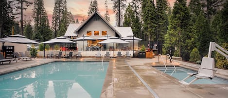 Un área de piscina con sillas de descanso y sombrillas, un edificio con techo inclinado y un fondo de bosque.