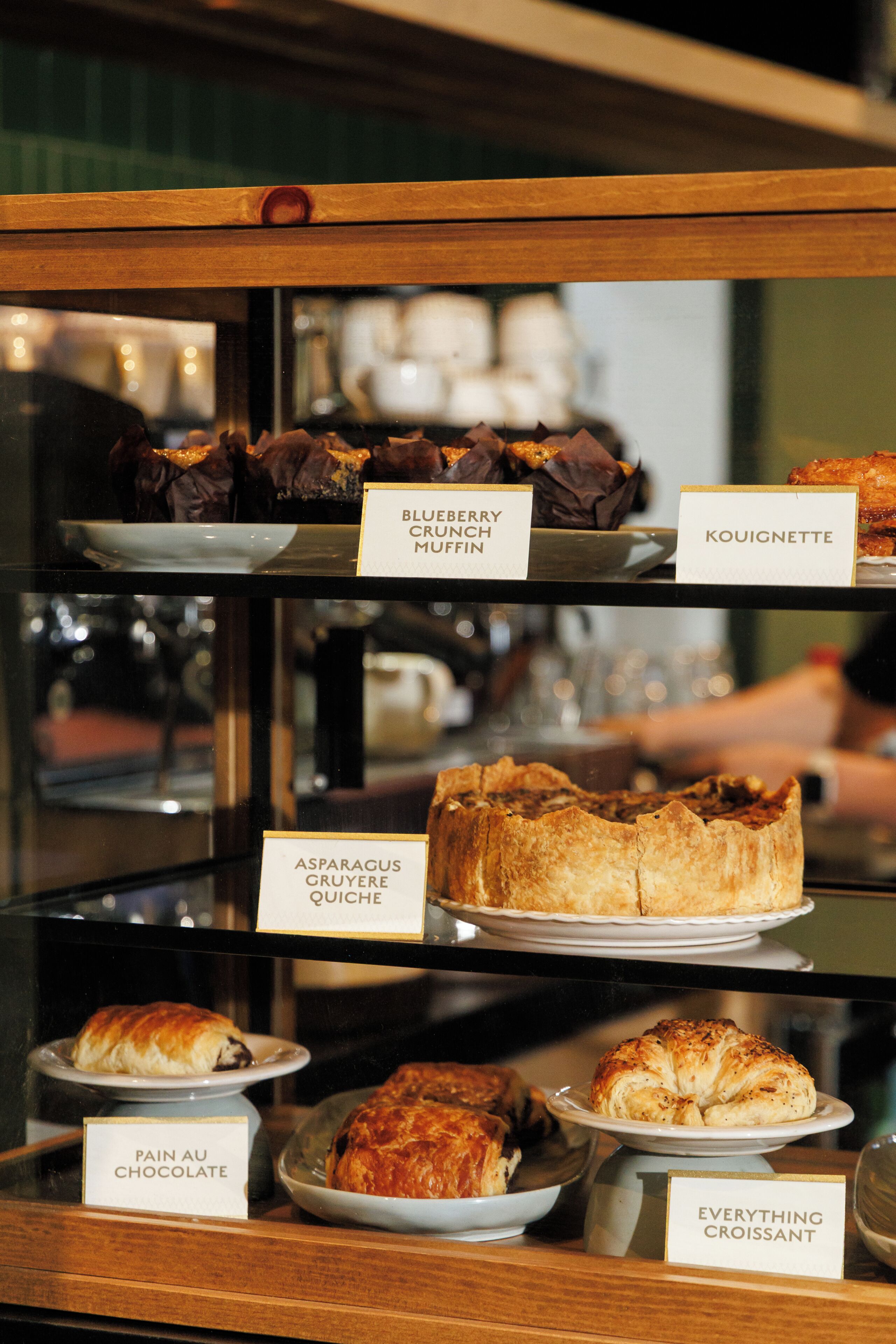 A display case with various baked goods labeled, including 'Blueberry Crunch Muffin' and 'Asparagus Gruyere Quiche'.