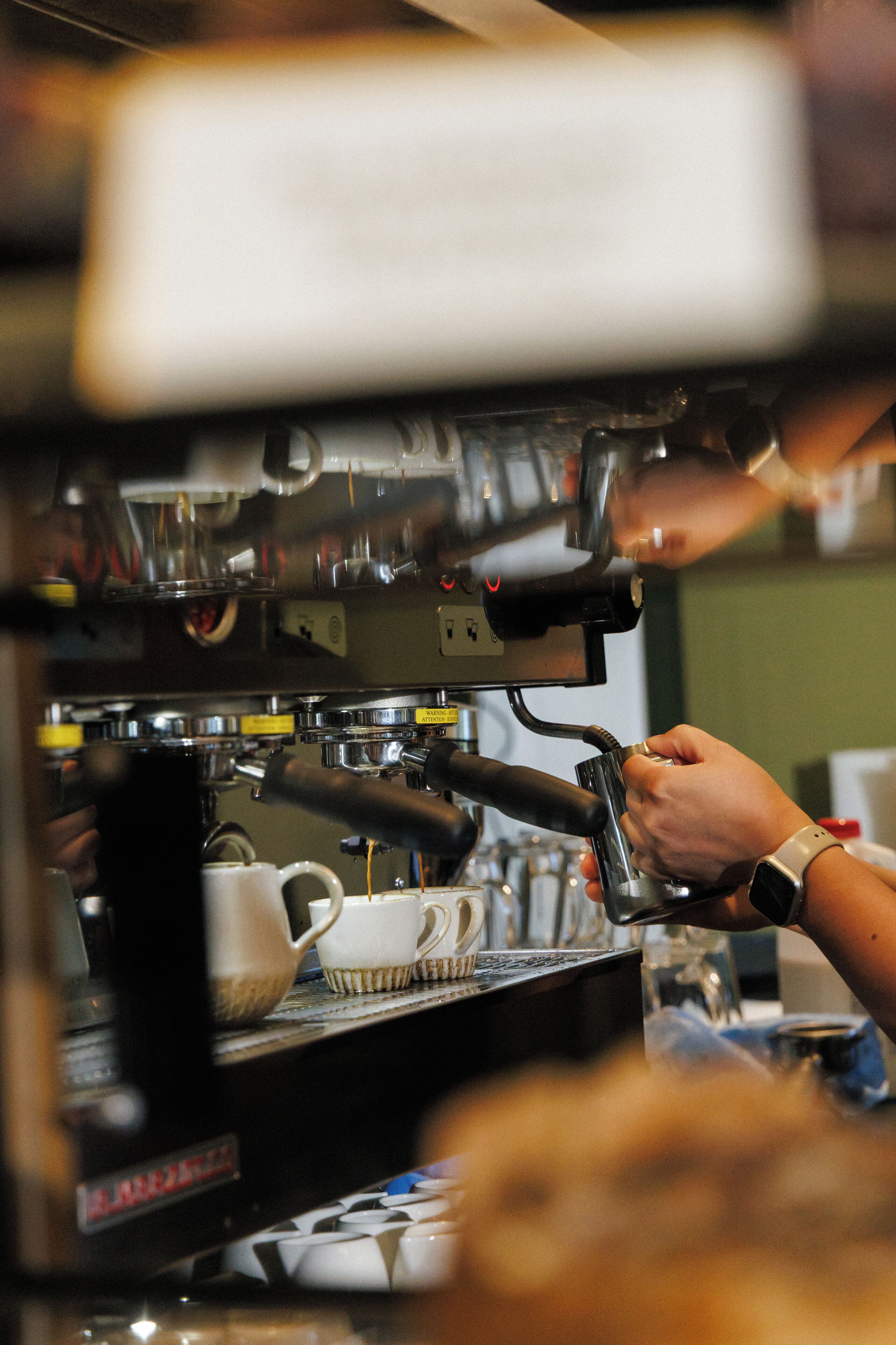 Une machine à café avec une personne qui l’utilise, des tasses et une montre au poignet.