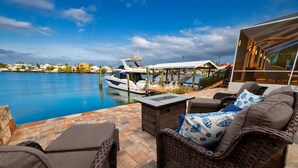 Um terraço à beira-mar com móveis de vime, piscina e um iate atracado por perto.