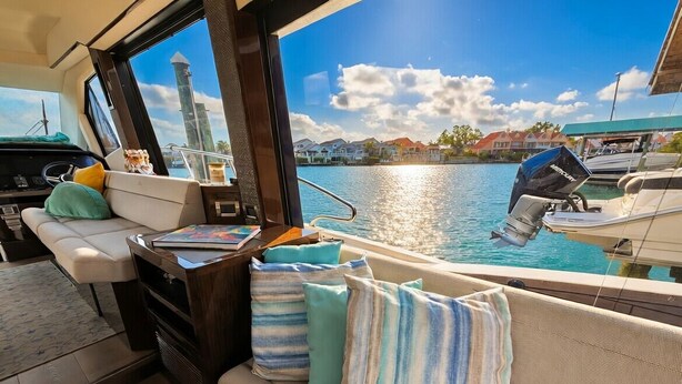 Interior de barco com área de estar acolchoada, painel de controle e vista para a água e casas.