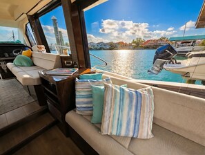 A boat interior with a cushioned seating area, a control panel, and a view of the water and houses.