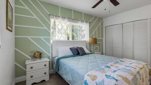 Ein Schlafzimmer mit Bett, Nachttisch, Kommode, Fenster mit Vorhängen und Deckenventilator.