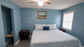 Une chambre à coucher comprenant un lit, une table de chevet, une petite table, un ventilateur de plafond, une fenêtre et un tableau accroché au mur.