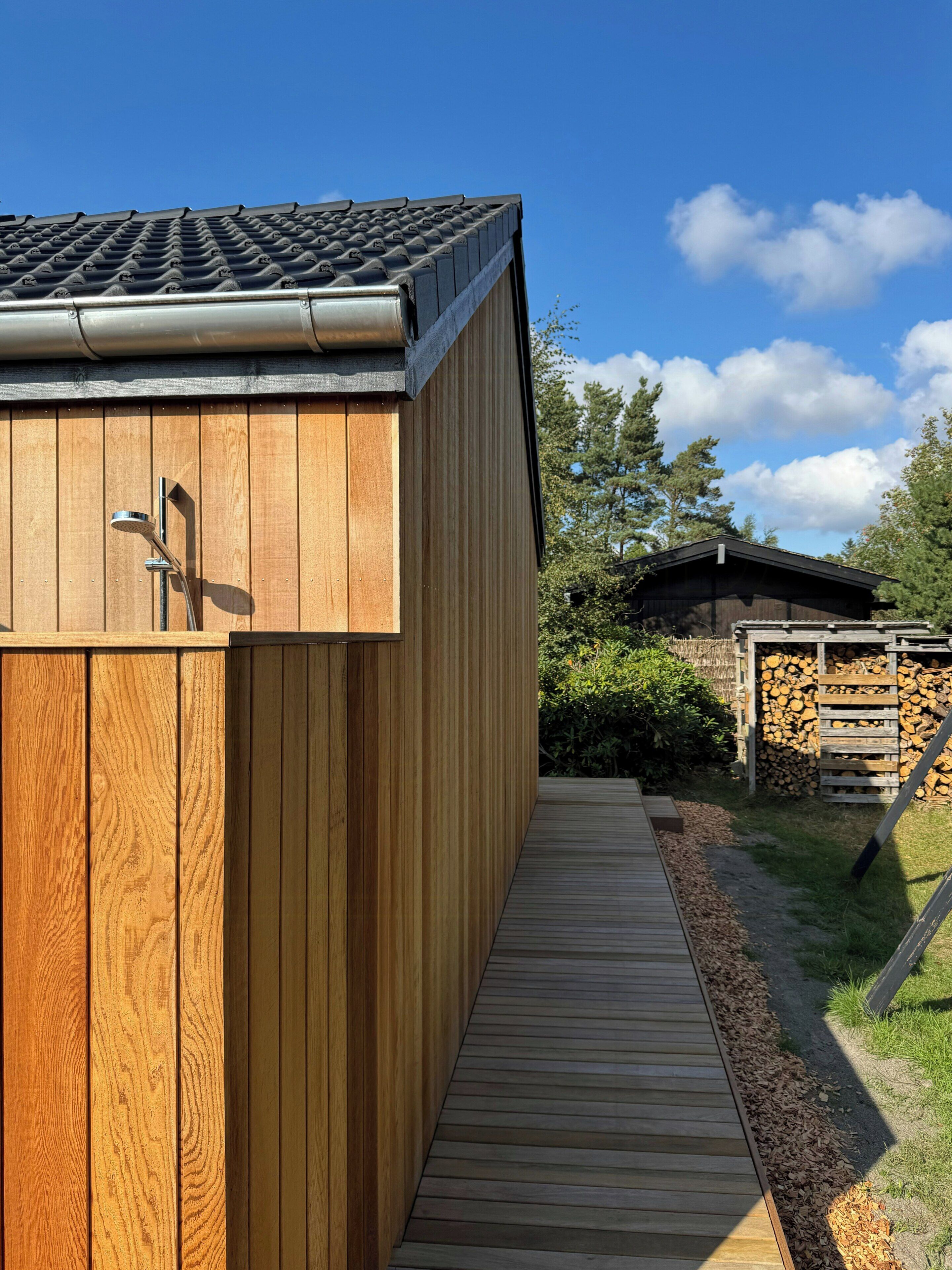 Een houten tuinhuisje met een metalen dak, een houten terras en een stapelhout op de achtergrond.