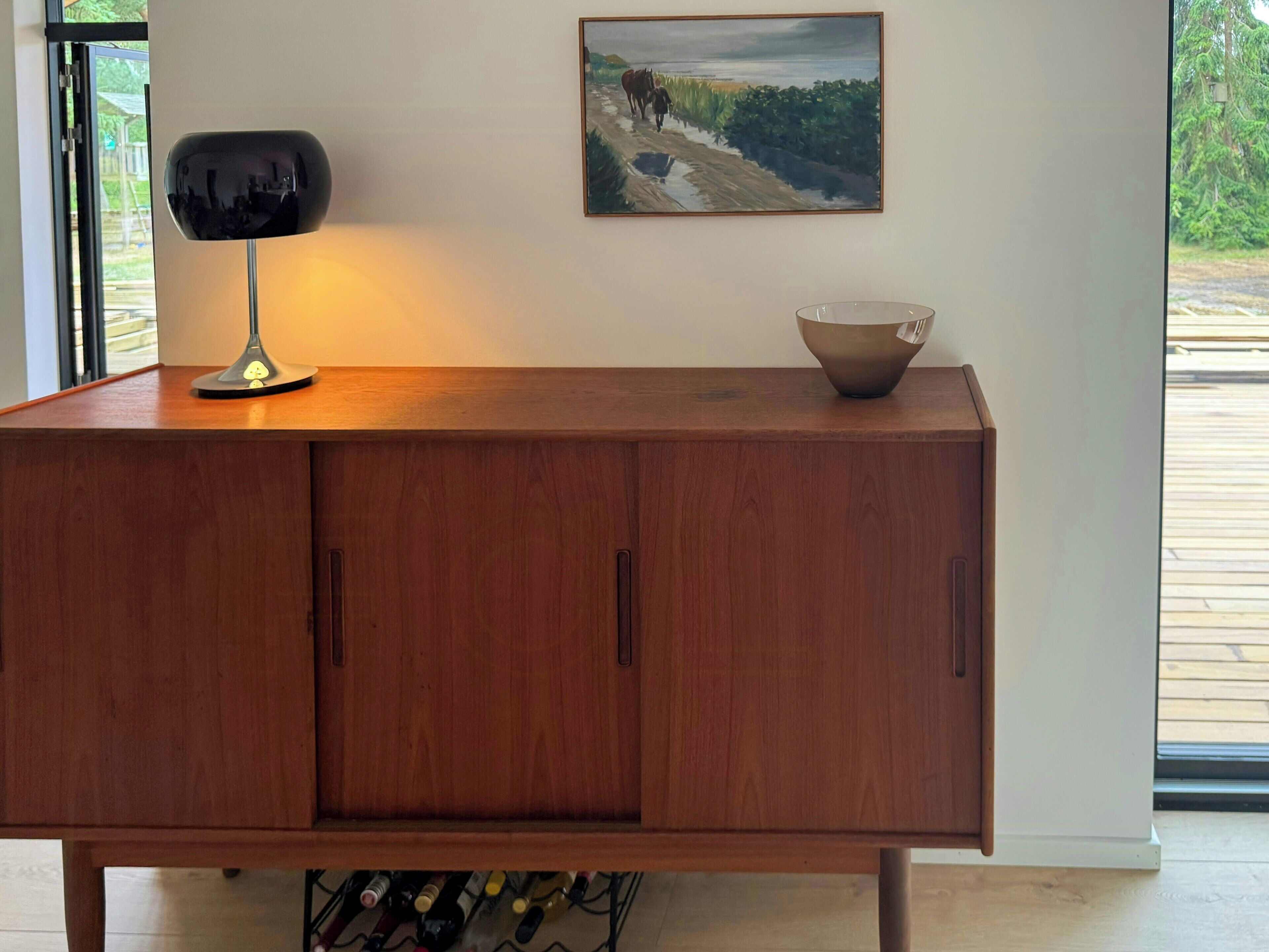 A wooden cabinet with a lamp and a bowl on top, a painting on the wall, and a view of greenery outside.