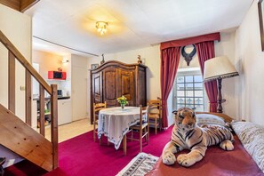 Una habitación con una cama, una silla de madera, una mesa con mantel, una lámpara, un peluche de tigre, un armario de madera y una escalera.