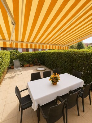 Patio esterno con una tenda a righe, un tavolo con sedie e un vaso di fiori.