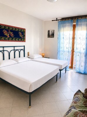 A room with two beds, a window with blue curtains, a painting on the wall, and a small table with a lamp.