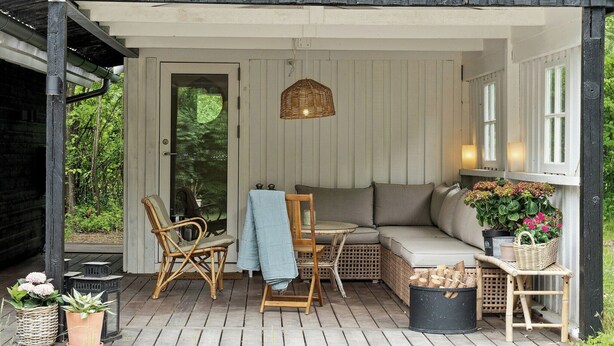 En overdækket veranda med siddeplads, en fletstol, et bord og en kurv med blomster.