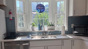 A kitchen with white cabinets, a stainless steel dishwasher, a double sink, and a window with a view of buildings and trees. - Midtown Gem Steps to Piedmont Park / Stylish 1BR (Atlanta)
