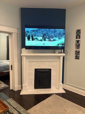 Una sala de estar con chimenea, un televisor montado encima y una cama al fondo.